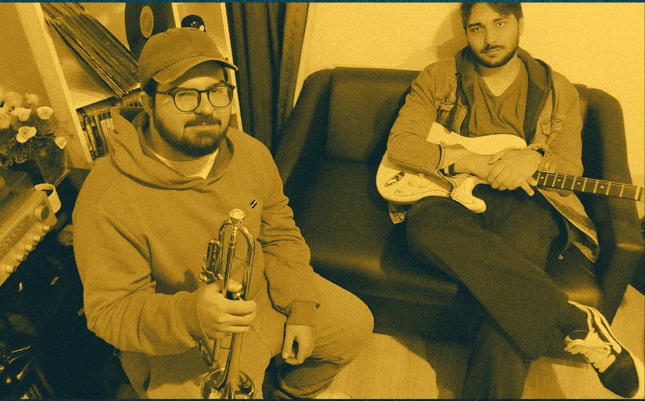 Os dois integrantes do Hotel Brasil sentados num sofá, um com o trompete e o outro com a guitarra